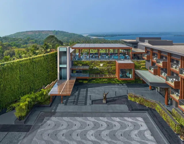 A modern hotel complex is shown above landscaped grounds with a coastal bay and green hills beyond.