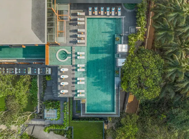 An aerial view shows a large pool surrounded by loungers, landscaped gardens, and tropical palm trees.