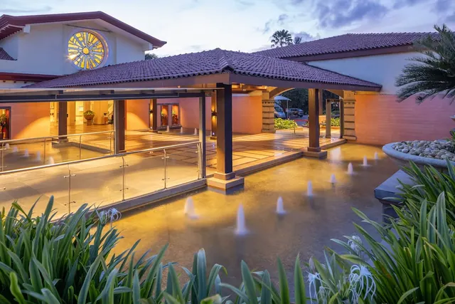 A covered hotel entrance with tiled roofs, illuminated windows, fountains and tropical landscaping is shown at dusk.
