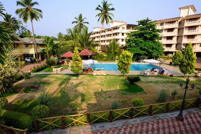 A landscaped hotel garden with a swimming pool, loungers, umbrellas, palm trees and surrounding resort buildings is visible.