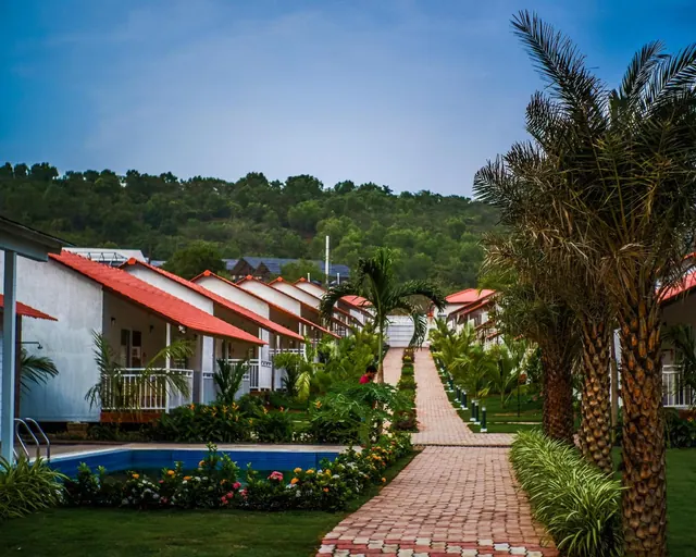 A landscaped resort garden has rows of white cottages, a brick walkway, palm trees and a small pool.