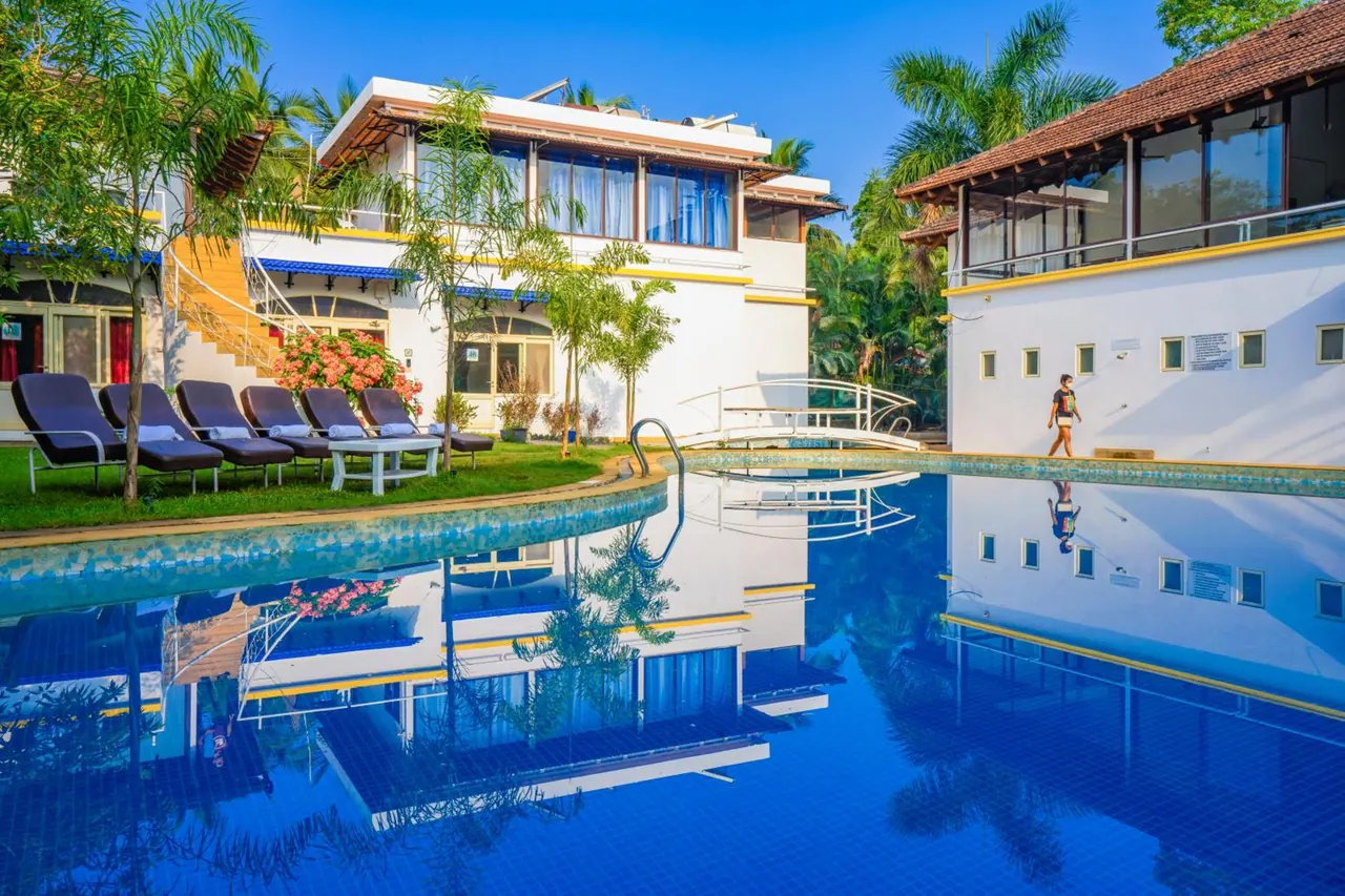 A large reflective swimming pool sits among sun loungers, tropical plants and white hotel buildings.