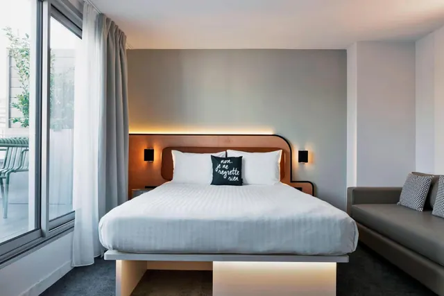 A modern hotel bedroom has a large bed, a sofa, and floor-to-ceiling windows opening onto a furnished balcony with a partial city view.