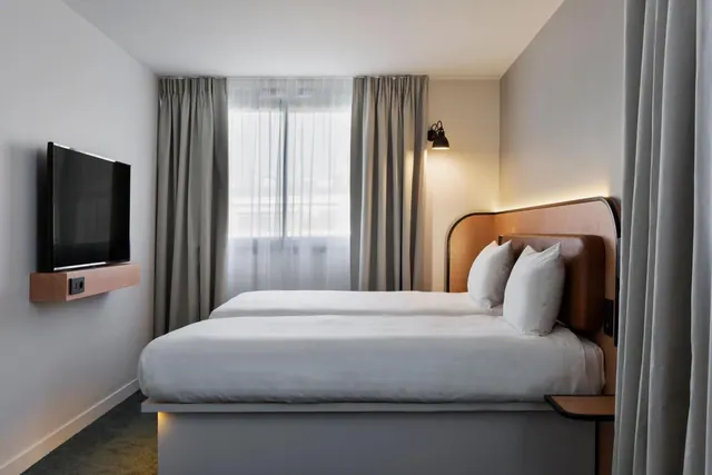 A modern hotel bedroom contains two twin beds, a wall-mounted television, and a curtained window.