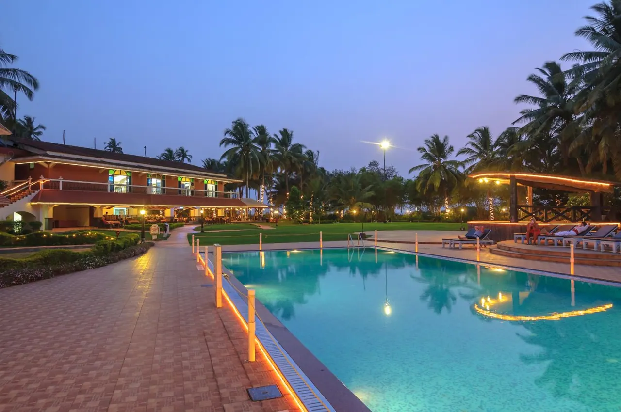 A large illuminated pool overlooks landscaped grounds and palm trees beside the hotel buildings at dusk.