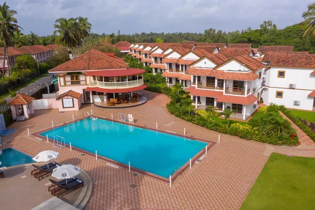 A large swimming pool and sun loungers sit among white buildings with red tiled roofs and tropical gardens.