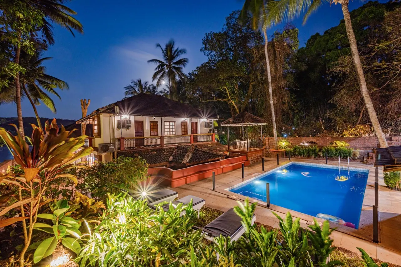 A lit swimming pool sits beside a traditional tiled-roof building surrounded by tropical gardens and palm trees.