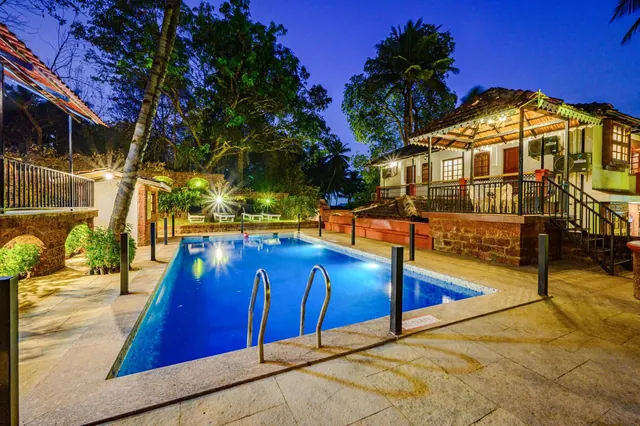 A lit swimming pool sits among tropical trees and traditional tiled-roof buildings.