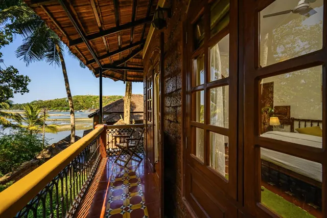 A furnished wooden balcony overlooks palm trees, tiled roofs and a waterway beside a heritage-style room.