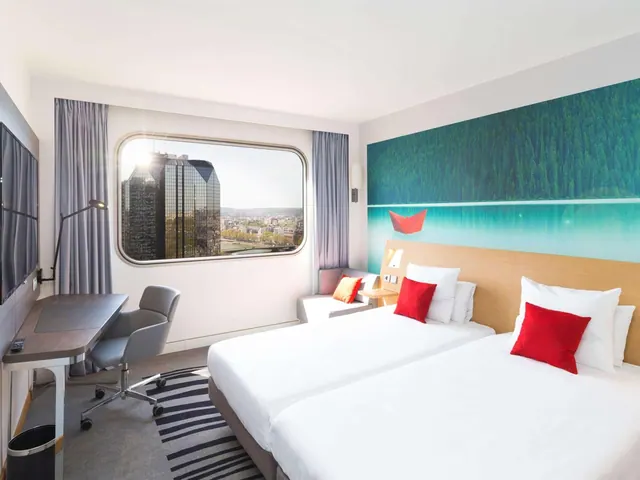 A bright modern hotel bedroom has twin beds, a desk, seating, and a clear city view through the large window.