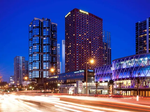 A Novotel hotel building and illuminated modern city buildings line a busy street at night.