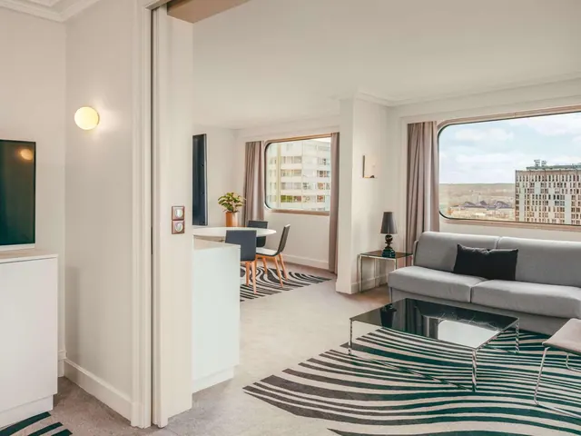 A bright living area has a sofa, television, dining table and large windows overlooking the city.