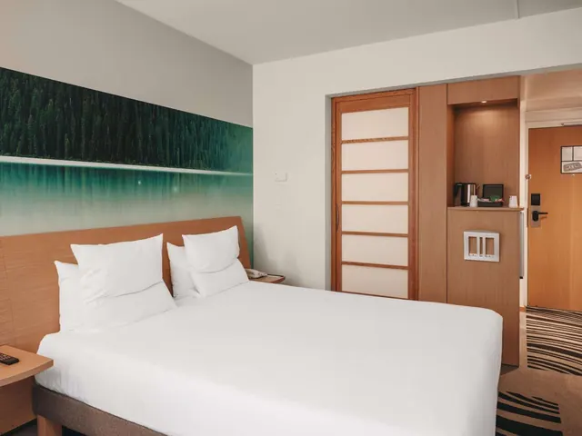 A bright modern hotel bedroom with a double bed, wood storage cabinetry and a coffee station.