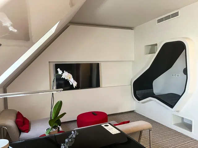 A modern hotel living area has sofas, a television, a desk, and a built-in cocoon chair beneath a sloped ceiling.