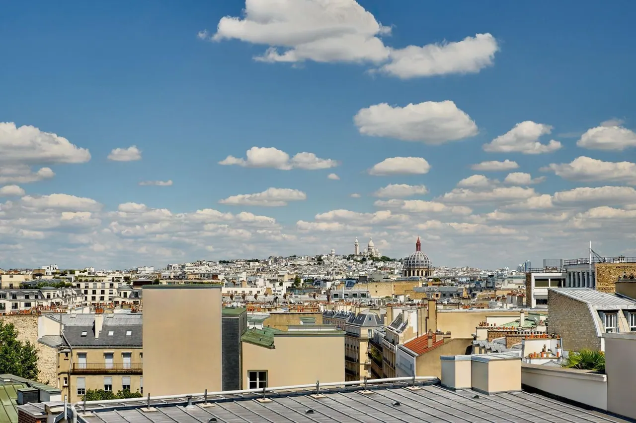 A broad Paris skyline view includes the Sacré-Cœur Basilica beneath a blue sky with scattered clouds.