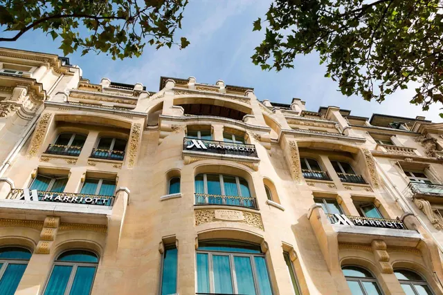 The ornate Marriott hotel facade is photographed from below beneath leafy tree branches.