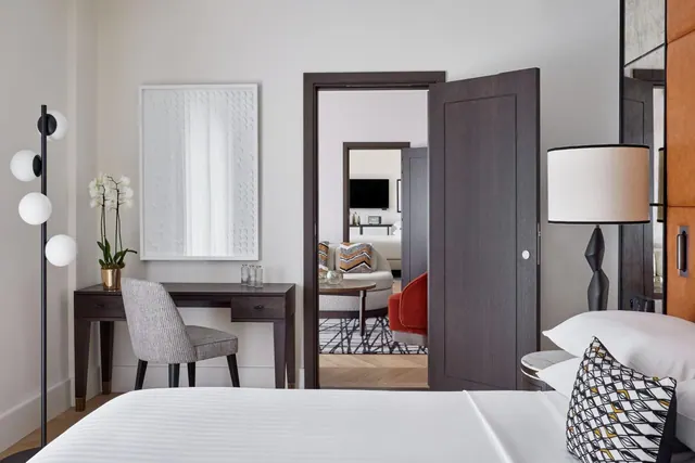 A bright modern bedroom has a neatly made bed, writing desk, decorative chair, lamp and mirrored doorway leading to a sitting area.