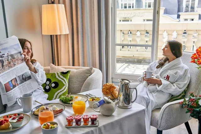 Two people in bathrobes sit in a sitting area beside a window while breakfast is served on a table.