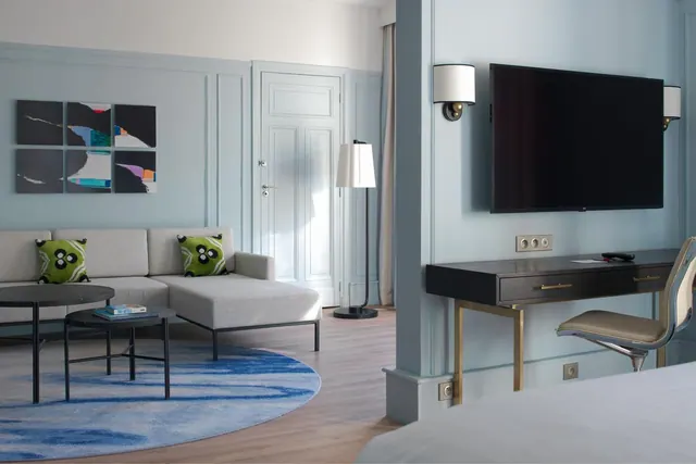 A bright hotel suite shows a sofa, coffee tables, desk, television, and part of a double bed.