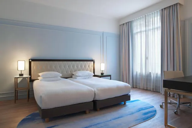 A bright hotel bedroom contains two separate beds, bedside lamps, a desk, and tall curtained windows.