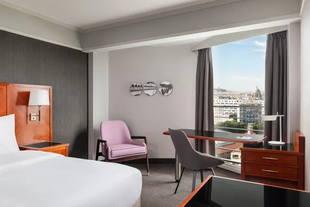 A hotel bedroom has a double bed, desk, armchairs and a broad city view through tall windows.