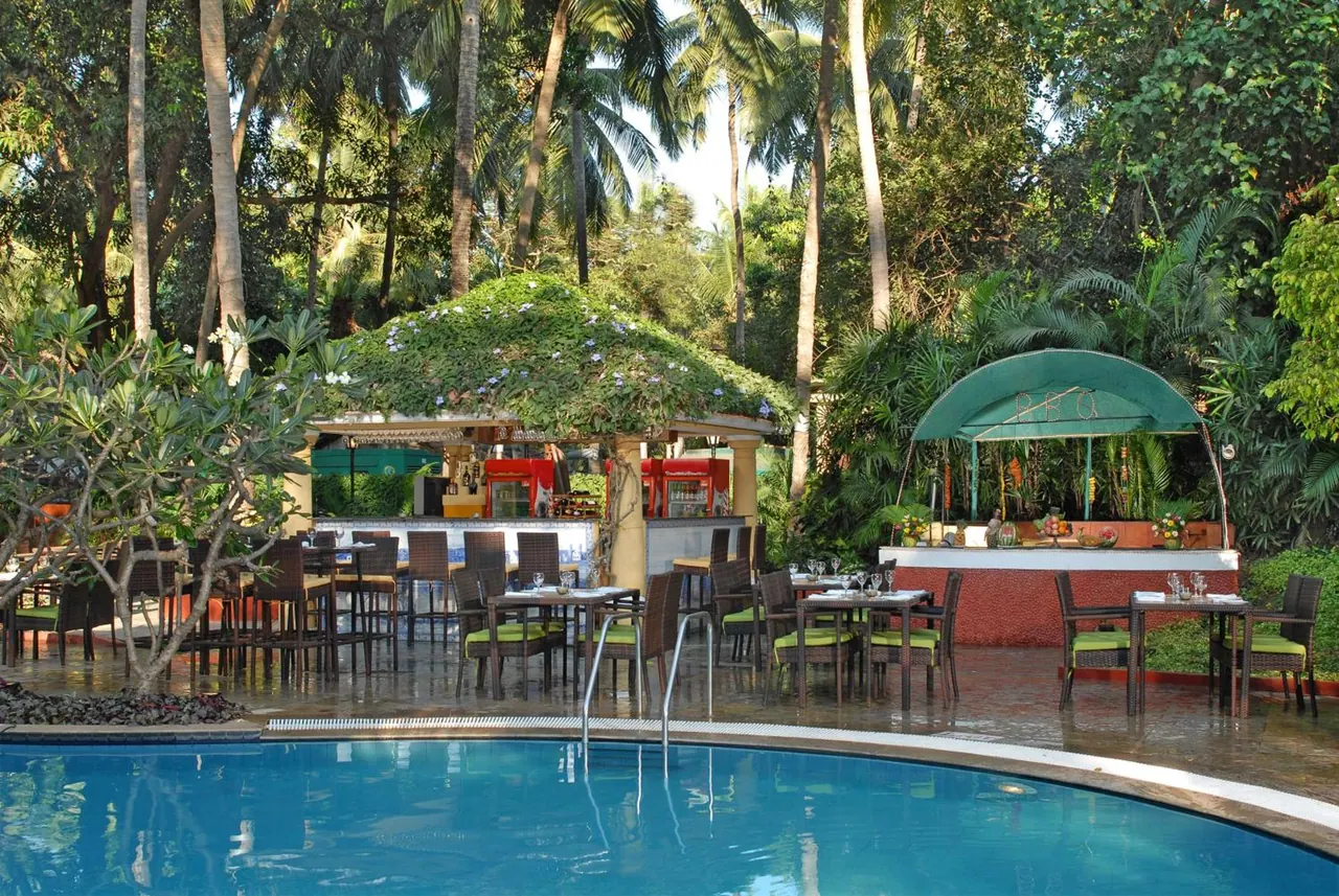 A large swimming pool is shown beside an outdoor dining area and pool bar surrounded by tropical vegetation.