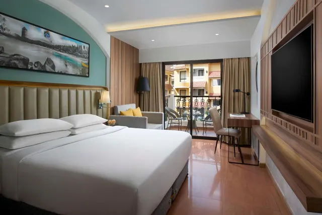 A bright modern bedroom has a double bed, seating area, desk, television and a furnished balcony overlooking surrounding buildings.