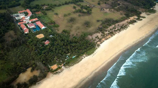 An aerial view shows a beachfront property surrounded by palm trees, fields and a sandy shoreline.
