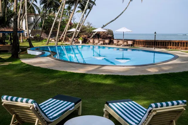 A large pool overlooks the sea beside a lawn, palm trees, loungers, umbrellas, and outdoor tables.