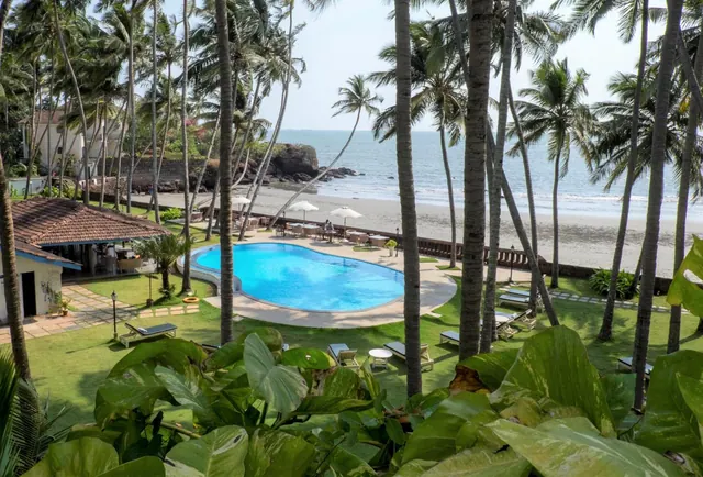 A tropical property overlooks a swimming pool, palm-filled grounds and a sandy beach beside the sea.