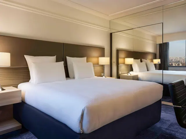 A bright hotel bedroom has a double bed, mirrored storage and a distant city view through the window.