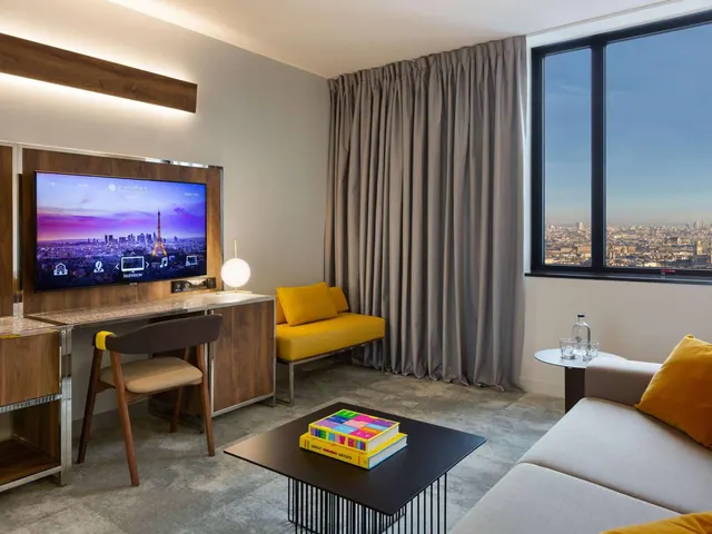A modern hotel living area has a desk, television, seating, and a clear city view through the window.