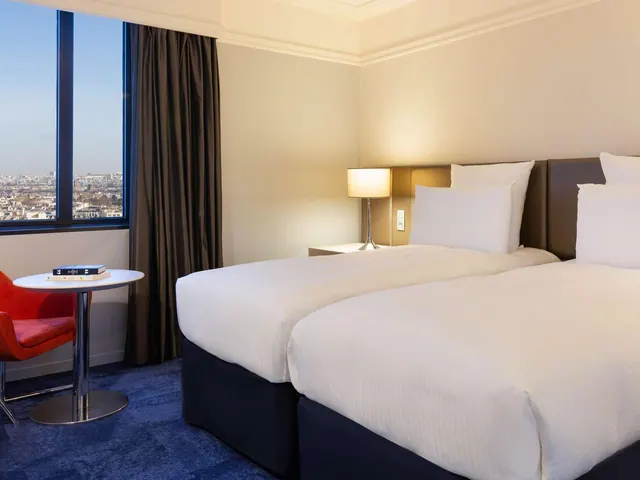 A bright hotel bedroom has twin beds, a small seating area, and a city view through the window.