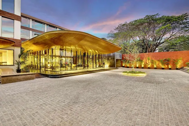 A modern hotel entrance with a broad curved canopy, glass walls, landscaped planters and a paved forecourt is shown at dusk.