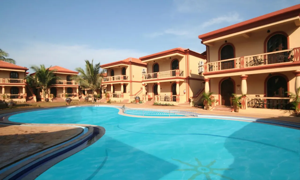 A large swimming pool fronts a tropical resort courtyard with two-storey buildings and palm trees.
