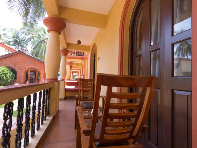 A furnished covered balcony overlooks a tropical garden and a red-brick building with arched windows.
