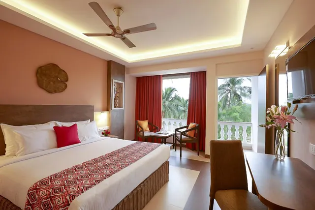 A bright bedroom contains a double bed, desk, seating area, ceiling fan and a balcony overlooking palm trees.