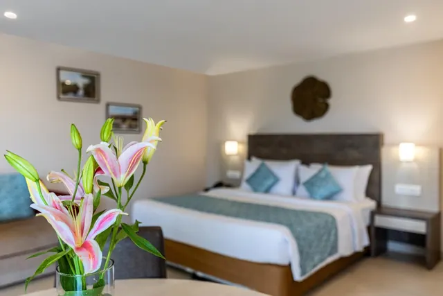 A neatly made double bed with blue accent pillows and a vase of pink lilies is shown in the bedroom.