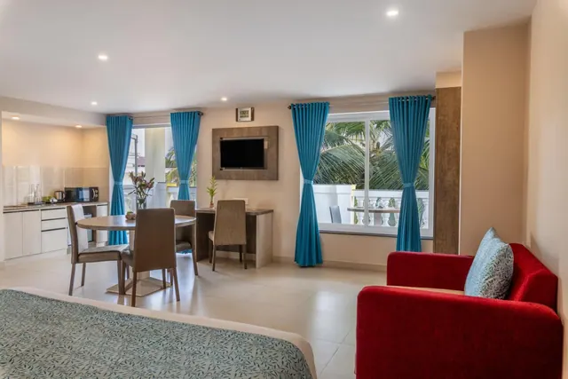 A bright hotel suite contains a bed, red sofa, dining table, kitchenette, television and windows overlooking palm trees.