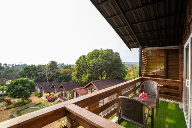 A furnished wooden balcony overlooks landscaped grounds, cottages, trees and distant hills.