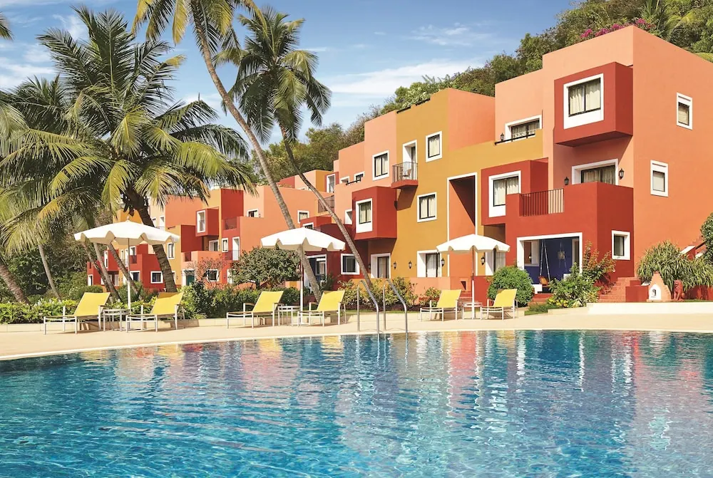 A large swimming pool with loungers and umbrellas faces colorful tropical resort buildings.
