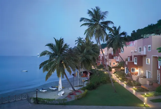 A palm-lined beachfront property overlooks the sea, sandy shore, boats, and landscaped grounds at dusk.