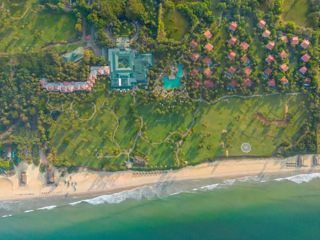 An aerial view shows a tropical resort, landscaped grounds, swimming pool and sandy beach beside the sea.