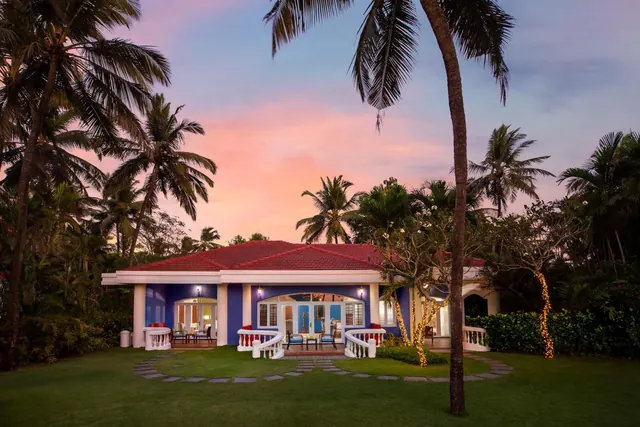 A brightly lit tropical villa stands amid palm trees and landscaped grounds at sunset.