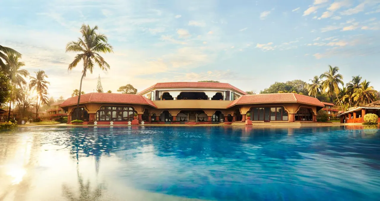 A large swimming pool reflects a tropical hotel building with red roofs and palm trees in warm evening light.