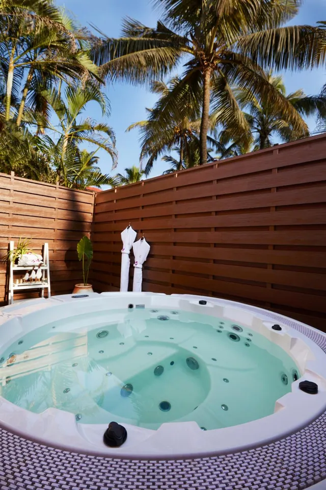 A private jacuzzi sits in a walled tropical courtyard with palm trees and two hanging robes.