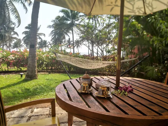 A wooden outdoor table with tea service sits beside a hammock overlooking a tropical garden.