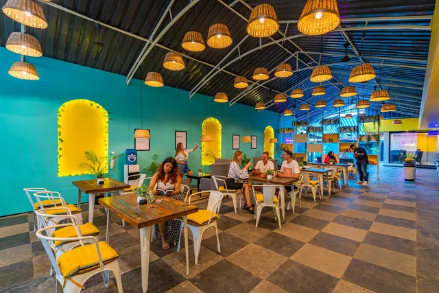 A colorful restaurant dining room has wooden tables, metal chairs, hanging wicker lamps and several diners.