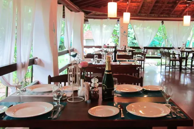 A covered dining room is arranged with set tables and views of green grounds through sheer curtains.