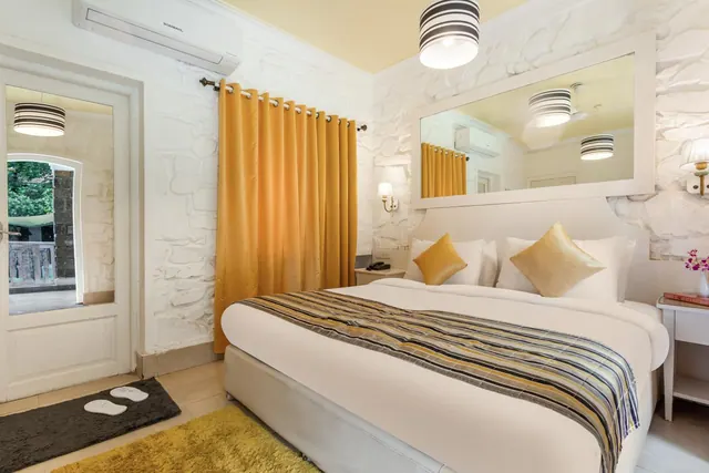 A bright bedroom with a double bed, patterned throw, yellow curtains, stone-textured walls and an air conditioner.